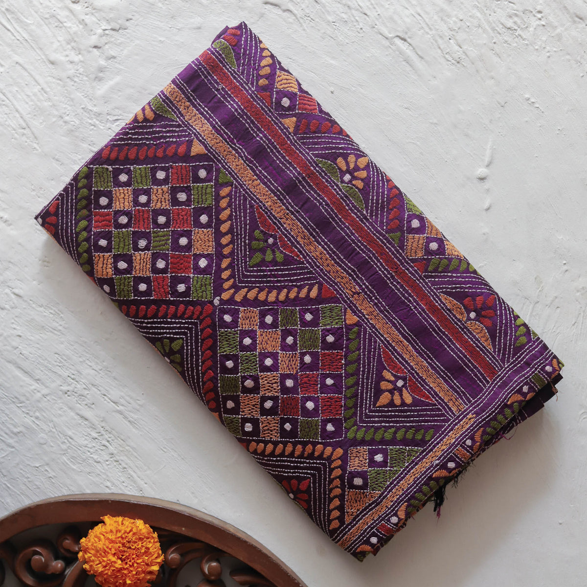 A folded Bengal handwoven mulberry silk hand embroidery kantha stitch saree of purple and multicolor patterned cotton fabric, likely a saree, displayed diagonally on a textured white wall with a decorative wooden bowl and orange flower in the lower left corner.
