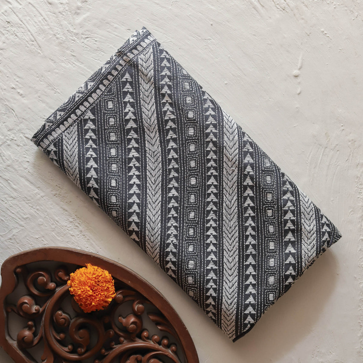 A Bengal handwoven mulberry silk hand embroidery kantha stitch saree with a black and white geometric tribal pattern, displayed diagonally on a textured off-white surface next to a decorative brown wooden tray with an orange flower.