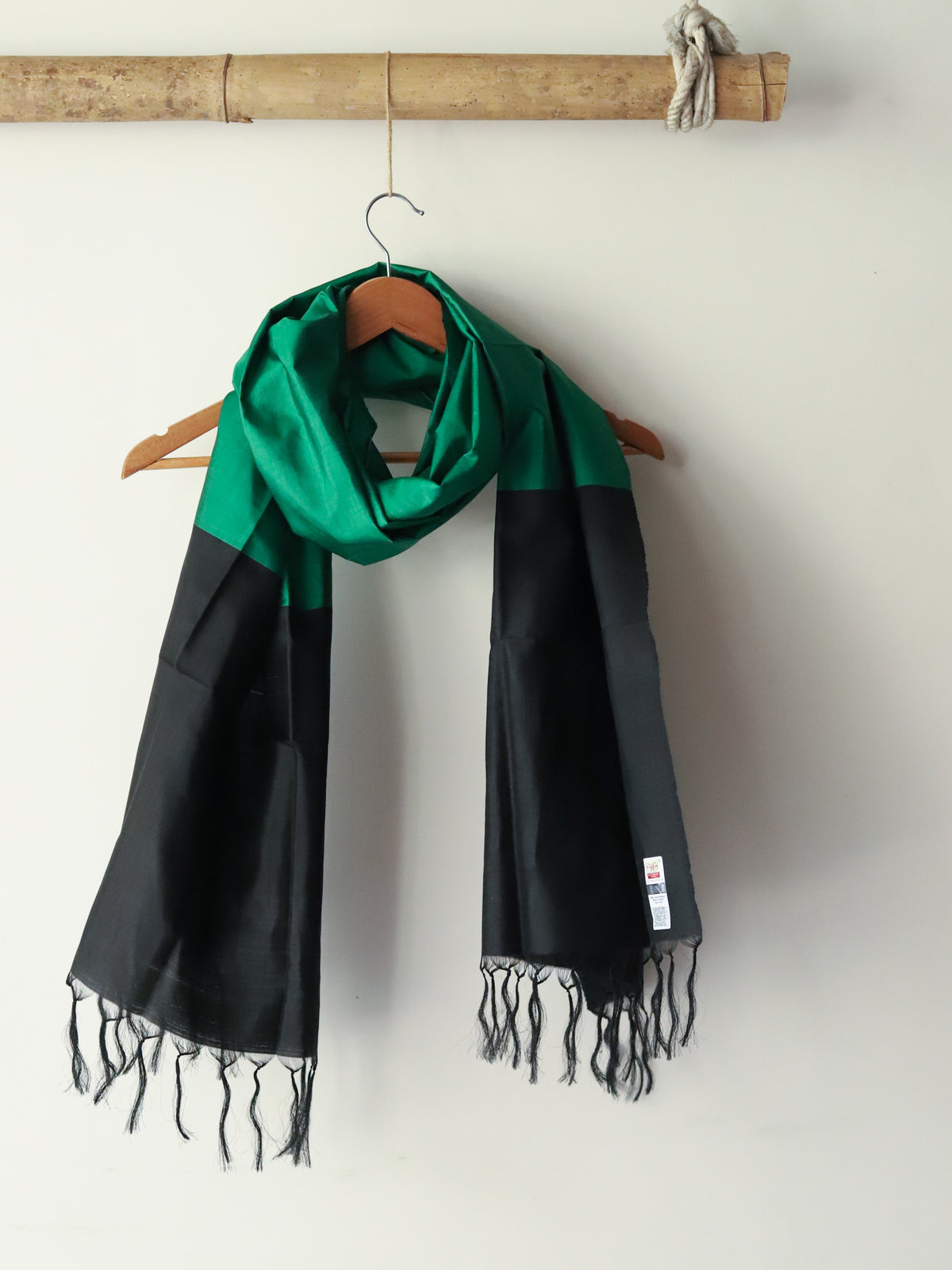 Green and black scarf with fringes hanging on a wooden hanger against a white wall.