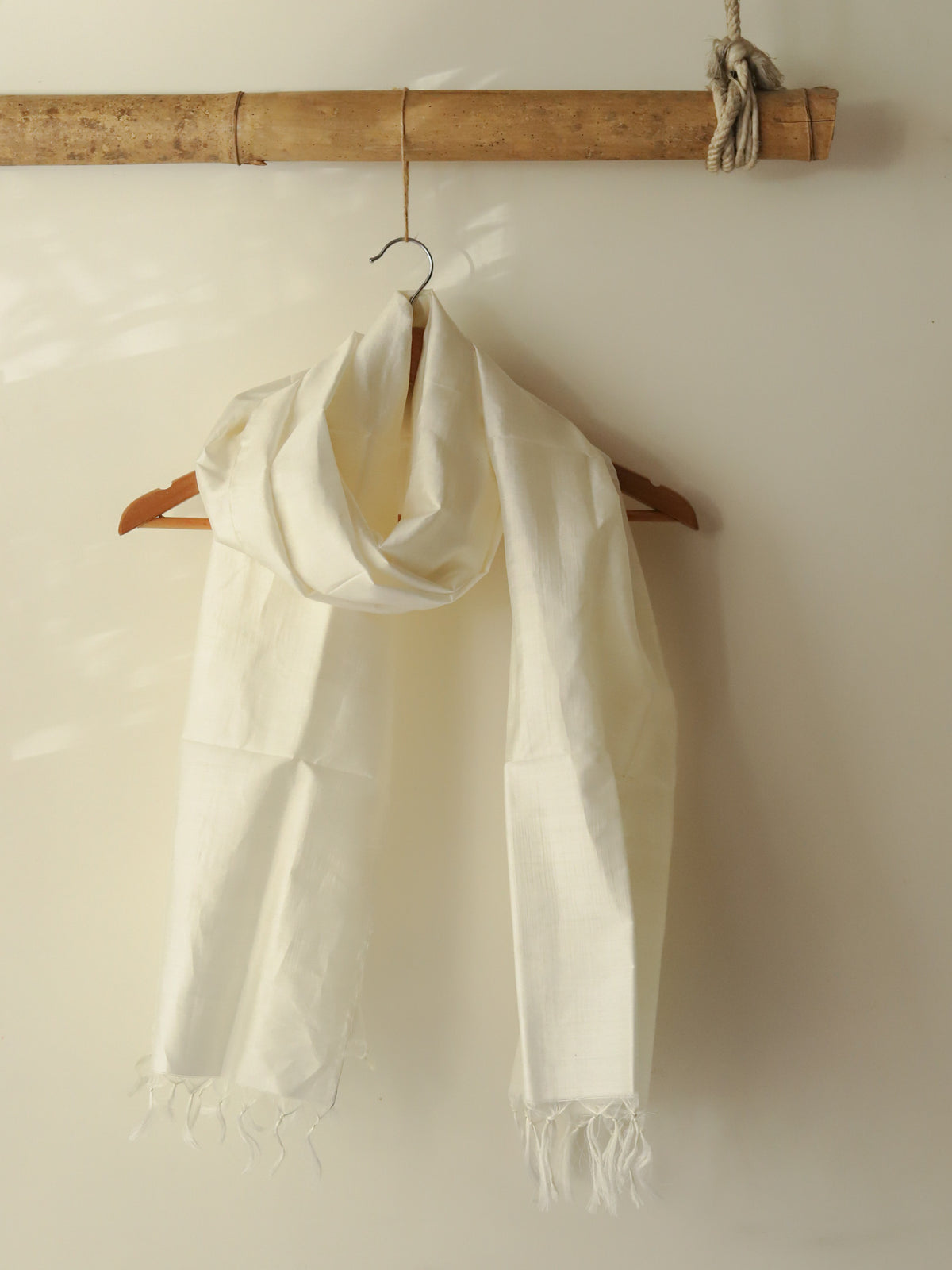 White scarf with fringes hanging on a wooden hanger against a white wall.