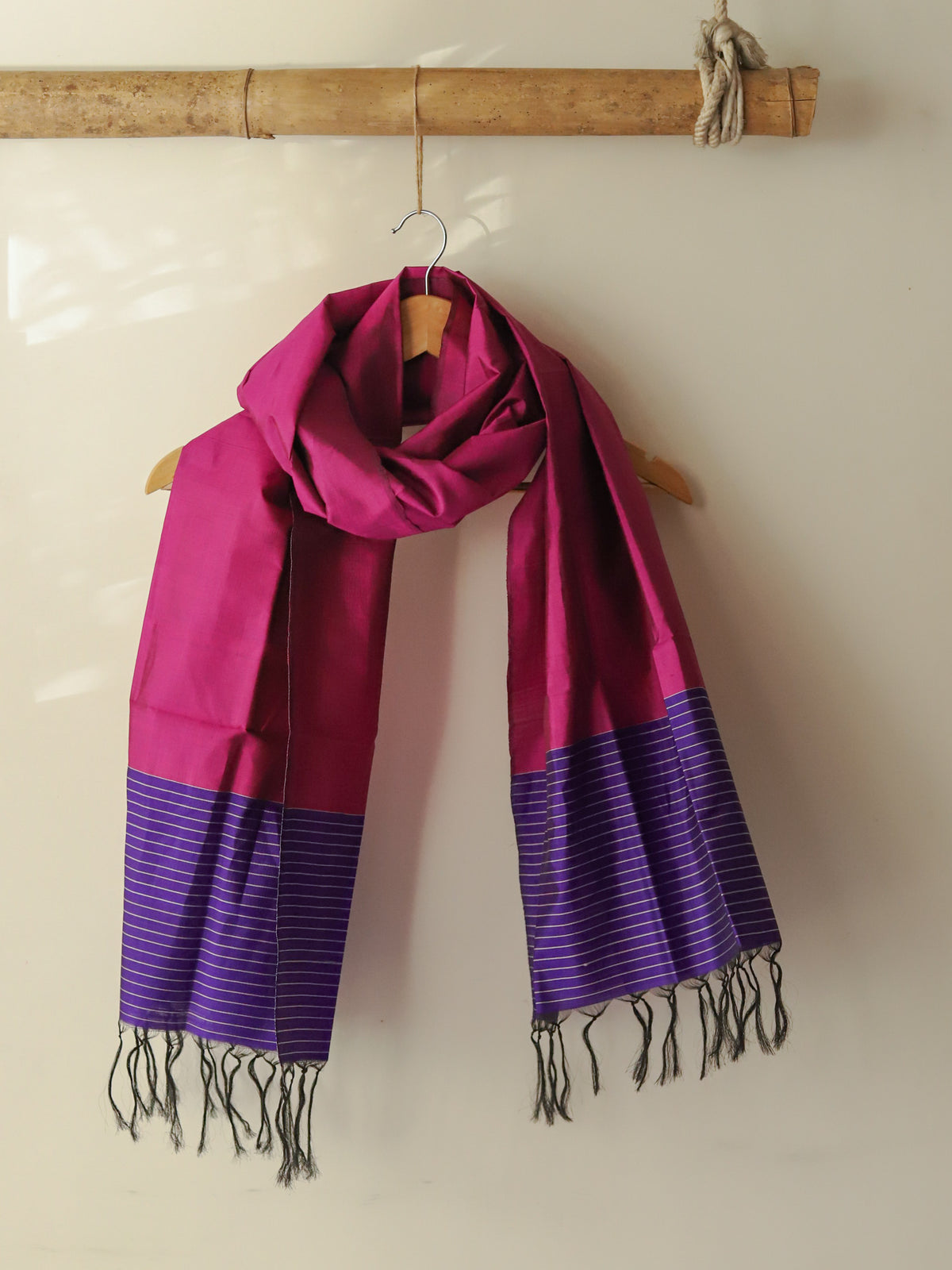 Pink and purple scarf with fringe on a wooden hanger against a beige wall.