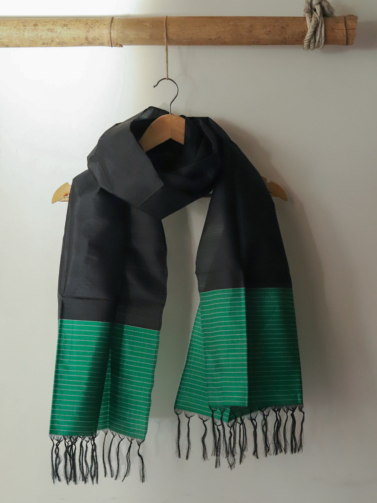 Black and green scarf with fringes hanging on a wooden hanger against a white wall.