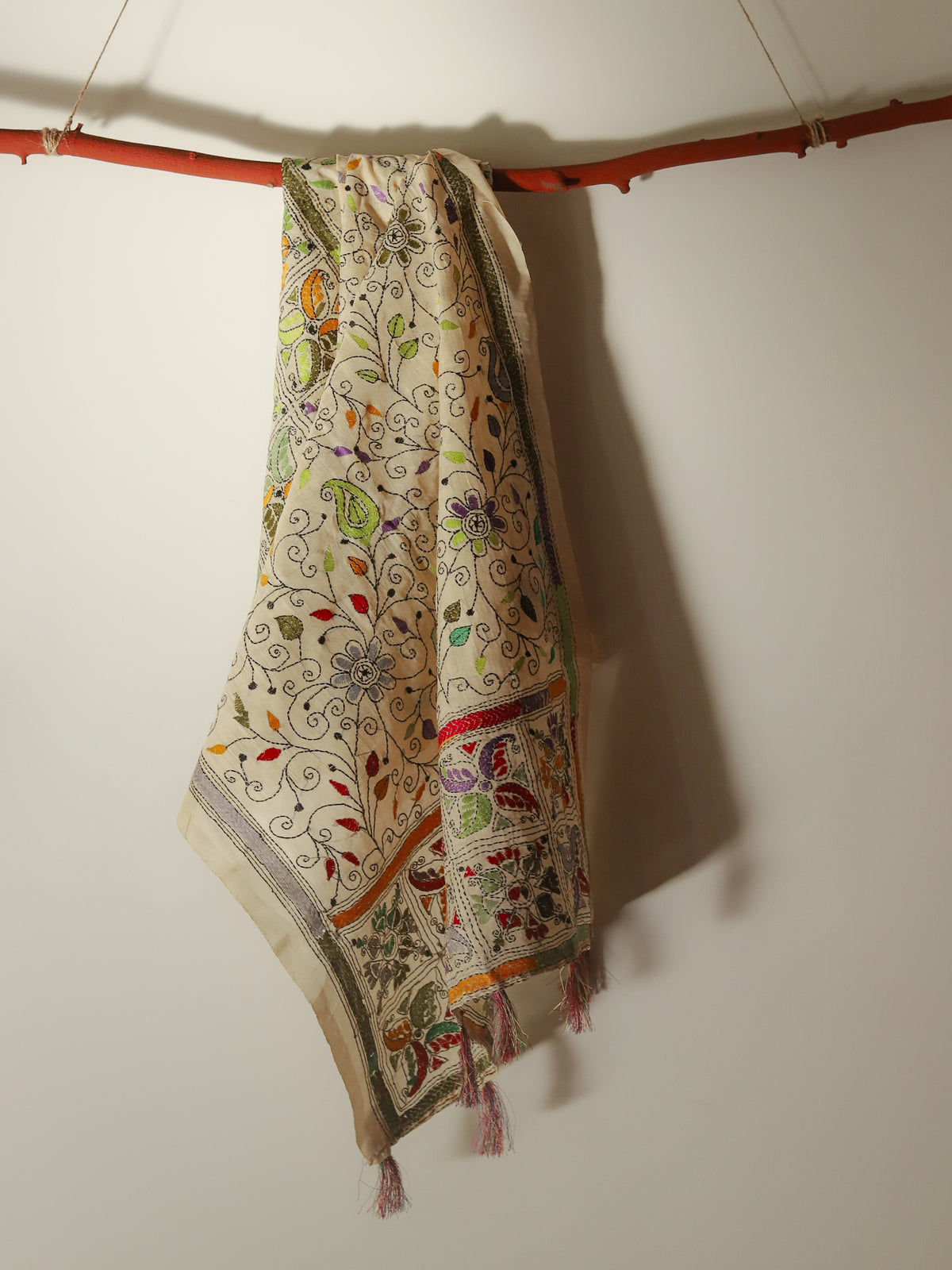 A cream-colored embroidered textile, Pobon Bengal Kantha Hand Embroidery Tussar Silk Stole or scarf or shawl, featuring intricate multicolor floral and paisley patterns, hanging from a rustic orange-brown wooden dowel against a plain off-white wall.
