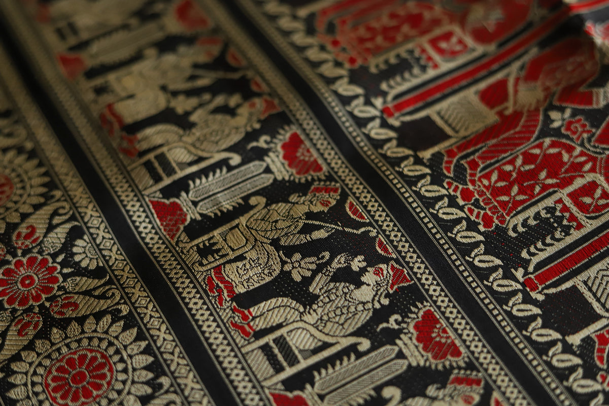 Close-up of intricately patterned fabric with red, black, and gold colors of Baluchari Saree.
