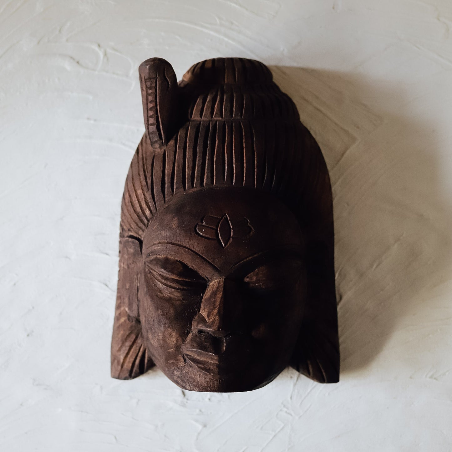 A hand-carved wooden mask with a traditional design, featuring intricate facial features, a forehead motif, and a hairstyle, displayed against a white background.