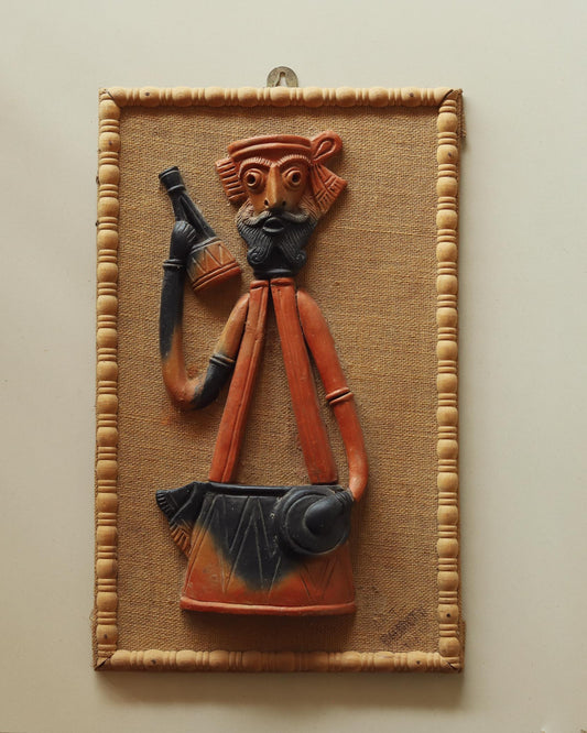 Handcrafted terracotta wall hanging depicting a Baul figure with miniature details, suspended on a jute board frame.