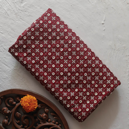 A folded rectangular Bengal hand embroidery kantha silk saree in maroon colour repeating white geometric block-print pattern, displayed on a textured off-white surface next to a carved dark brown wooden tray with an orange marigold.