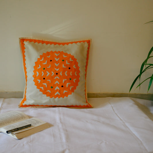 Handcrafted cushion cover with an orange and beige geometric pattern, placed on a couch next to a potted plant and a book.