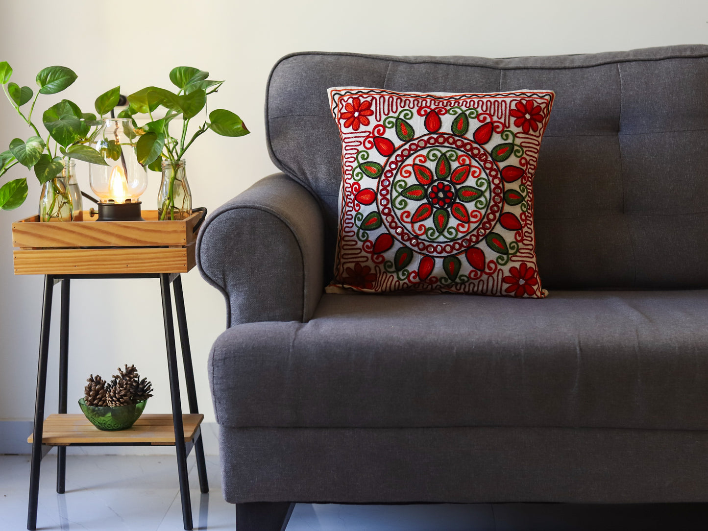 Gray sofa with a decorative pillow featuring red and green patterns, next to a small wooden side table with plants.
