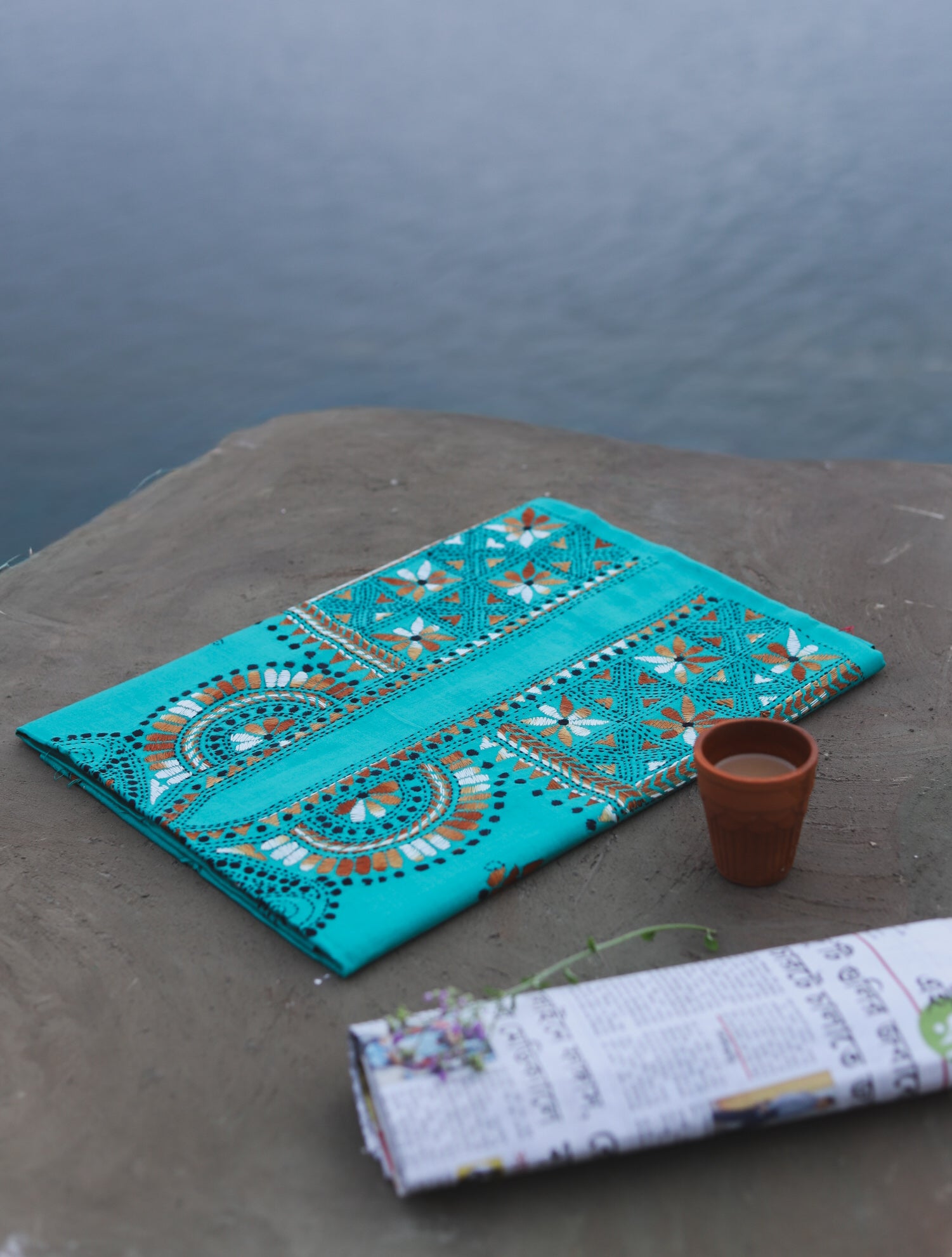 Turquoise embroidered fabric on a stone surface with a cup and newspaper in the foreground, against a water background.
