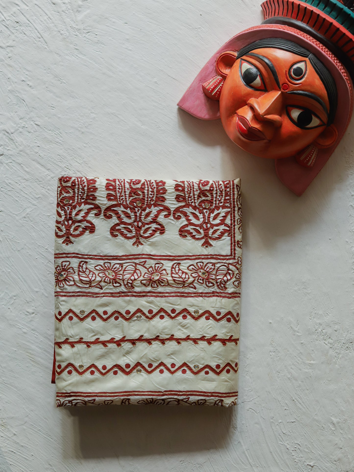 A folded Bengal handwoven red cream-colored mulberry silk saree with red hand-block printed patterns, displayed on a textured off-white wall next to a traditional orange and red folk art mask.