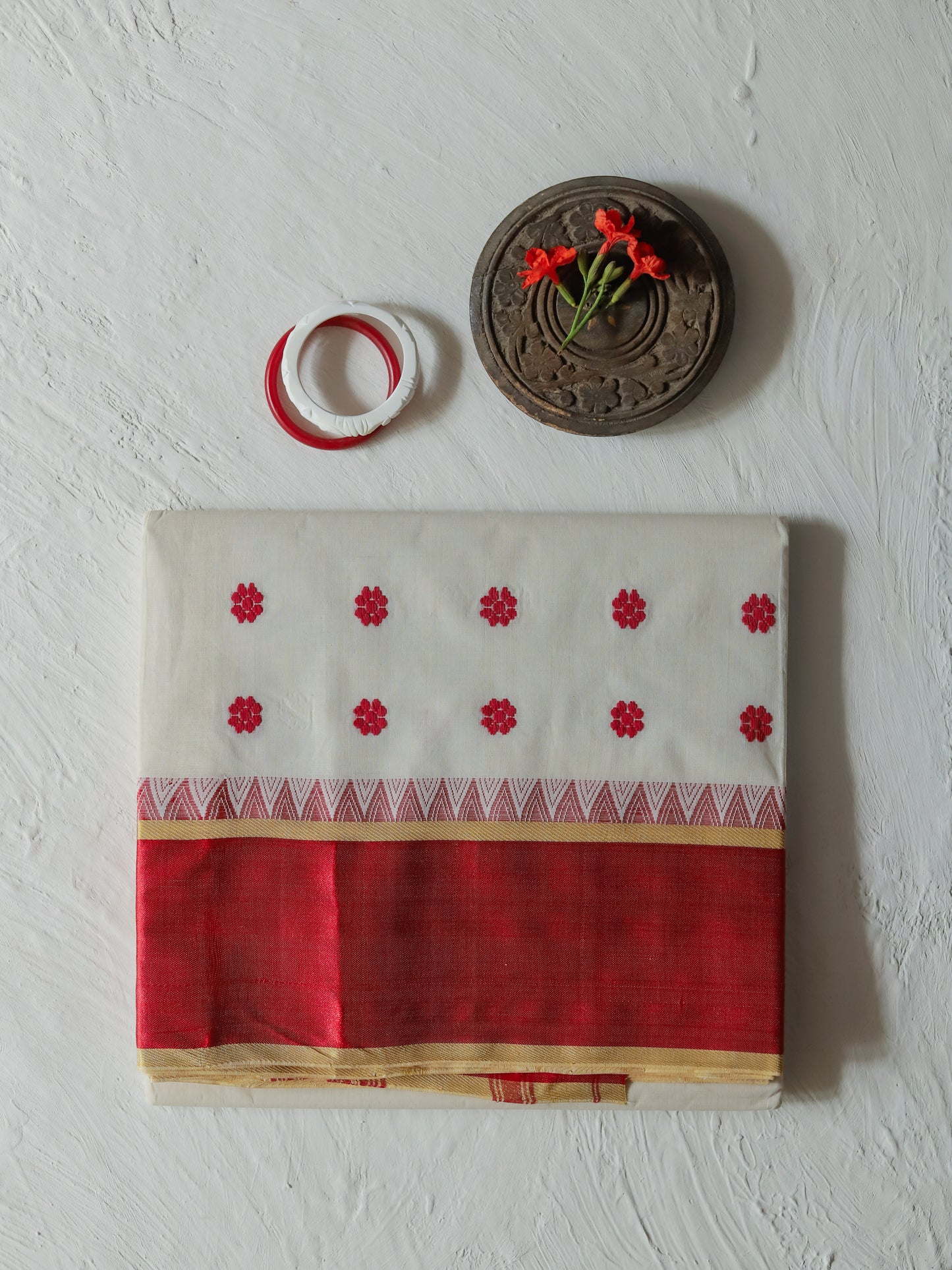 A folded Bengali red and white cotton saree with red floral motifs and a geometric border, displayed on a textured white wall alongside a small wooden decorative piece and a roll of red and white ribbon.