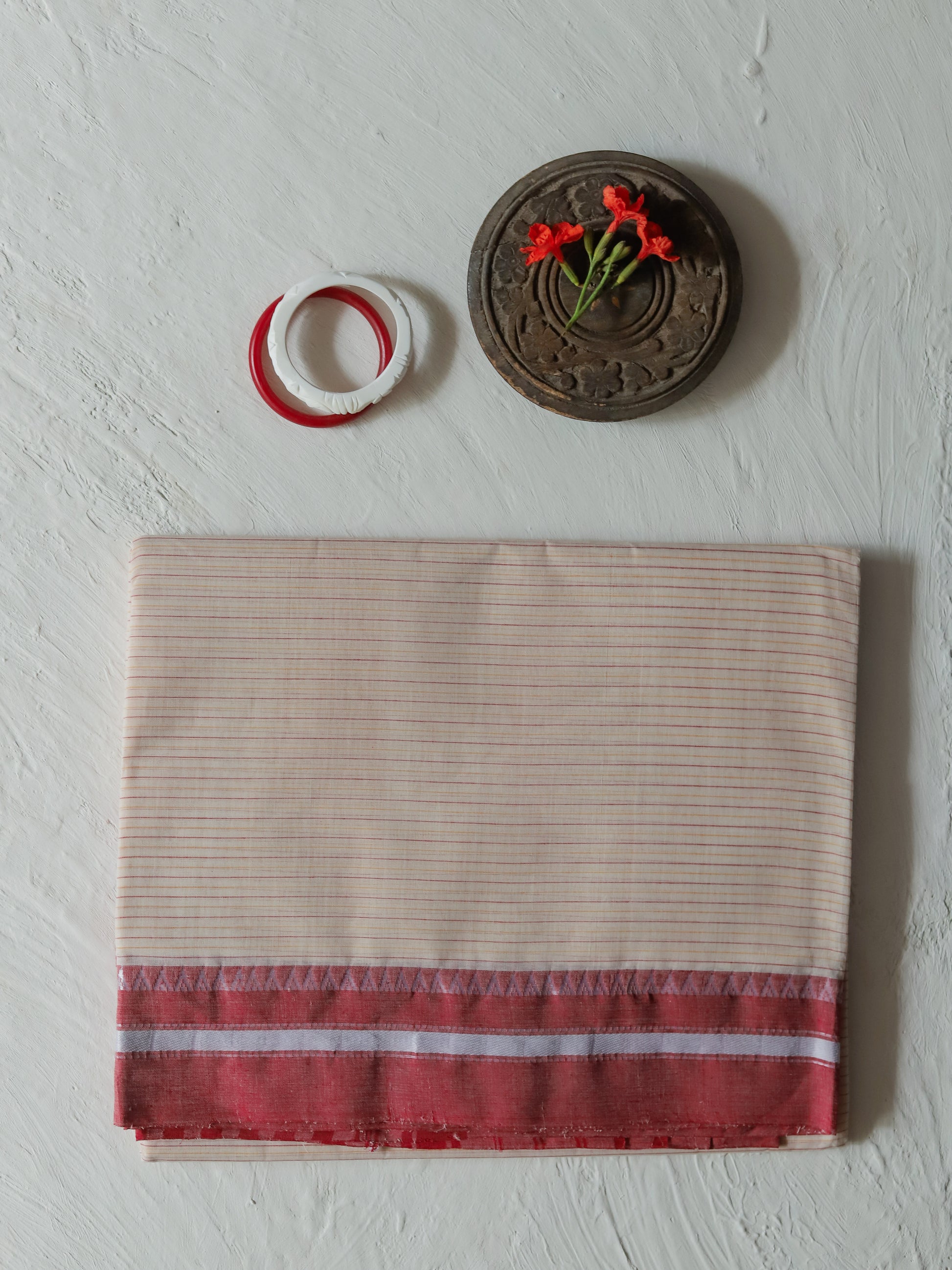 A folded Bengali white red cotton saree striped fabric with a red and white patterned border, displayed on a textured white wall alongside a small carved wooden decorative piece and a red-and-white ring.