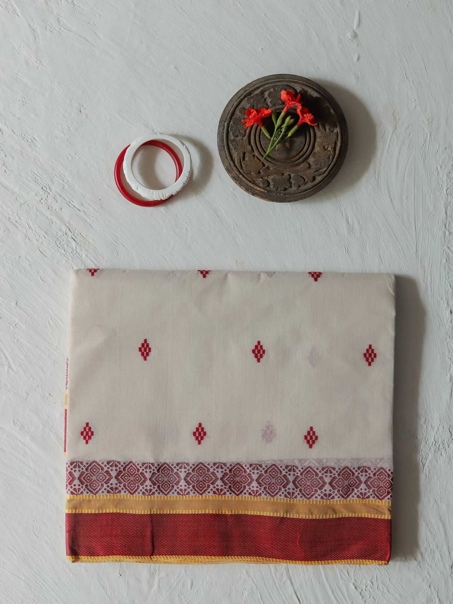 A folded Bengali handwoven white red cotton saree with red embroidered diamond motifs, a patterned border, and a red border, displayed on a textured white wall alongside decorative props.