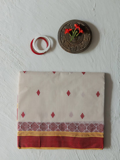 A folded Bengali handwoven white red cotton saree with red embroidered diamond motifs, a patterned border, and a red border, displayed on a textured white wall alongside decorative props.