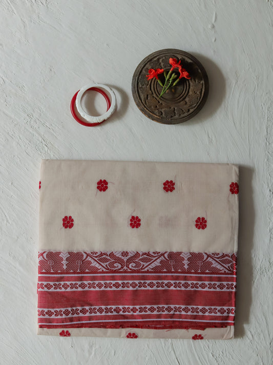 A folded colored Bengali white red cotton fabric piece, likely a sari or scarf, featuring red floral motifs and a wide red patterned border, displayed on a textured white wall alongside decorative props.