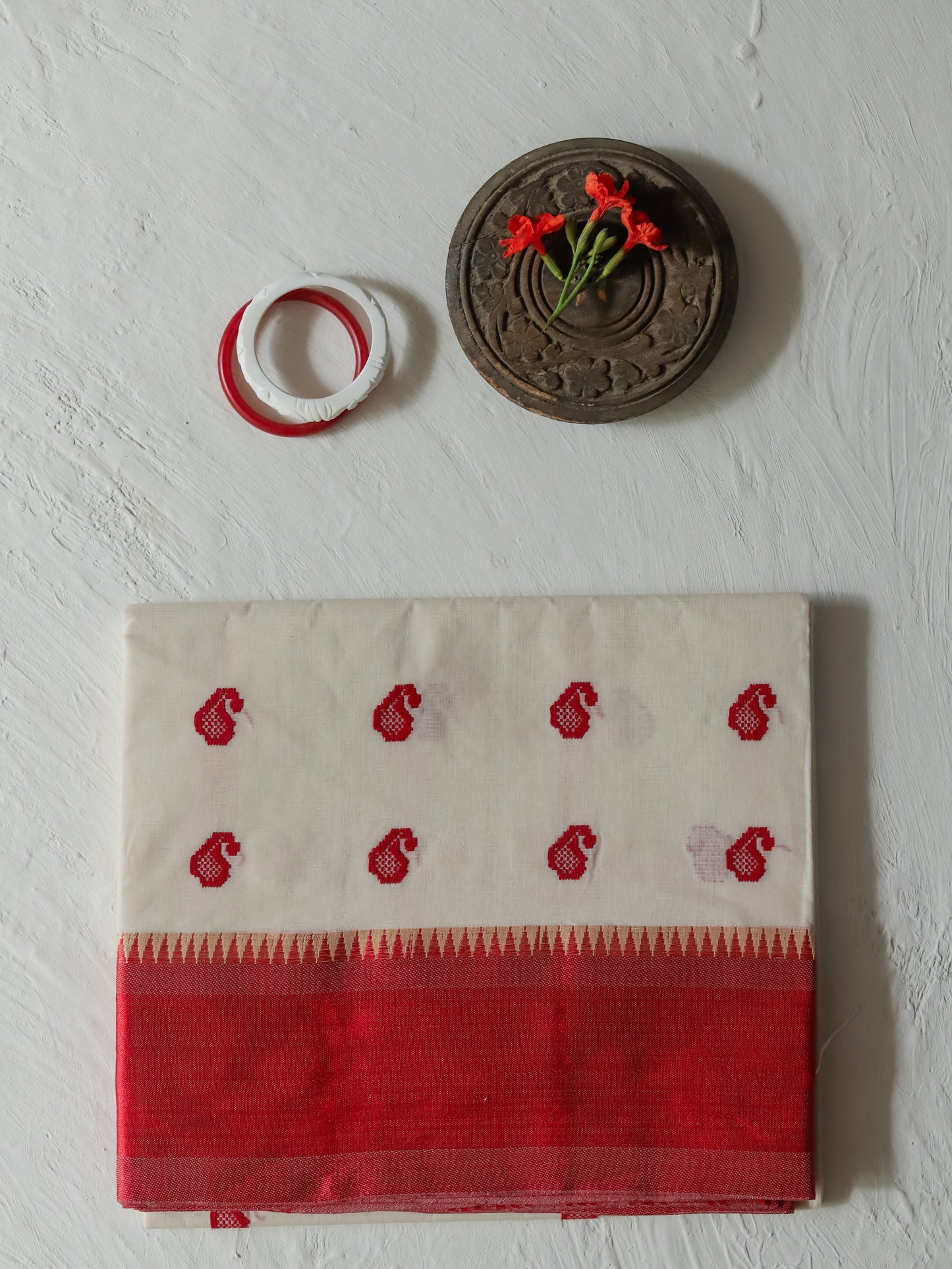 A folded Bengali red and white cotton saree or fabric piece with red handwoven motifs and a red border, displayed on a textured white wall below a small round wooden decor piece and a red-and-white ring.