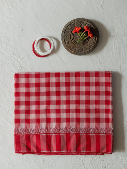 A folded Bengal red and white gamcha gingham checkered cotton saree with a ruffled border, displayed on a textured white background alongside a small decorative wooden piece and two white rings with red trim.