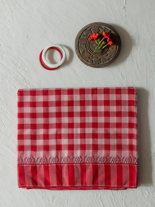 A folded Bengal red and white gamcha gingham checkered cotton saree with a ruffled border, displayed on a textured white background alongside a small decorative wooden piece and two white rings with red trim.