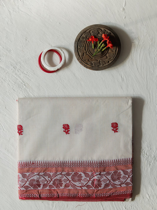A folded Bengali off-white cotton saree or fabric piece with red floral motifs and a decorative red border, displayed on a textured white wall alongside a small carved wooden ornament, red flowers, and two red-and-white rings.