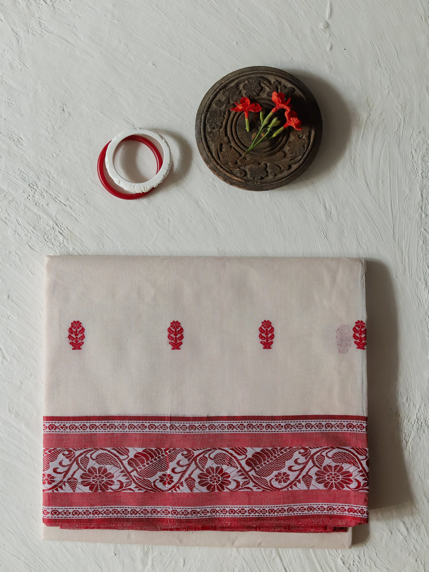 A folded Bengali red white cotton saree featuring red floral motifs and a wide red patterned border, displayed on a textured off-white wall alongside a small wooden decorative piece and a red-and-white ring.