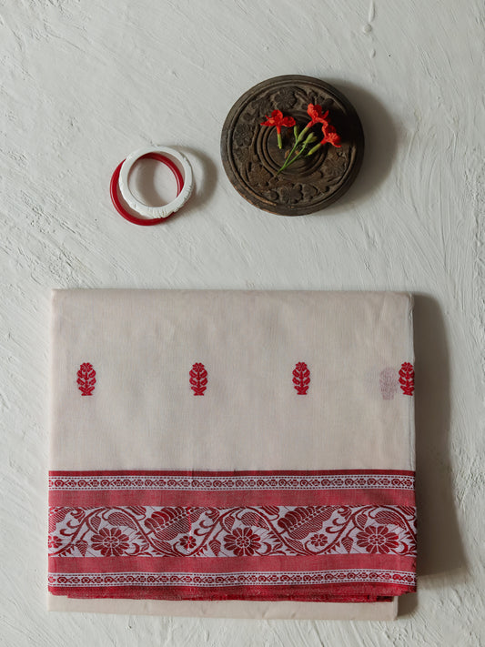 A folded Bengali red white cotton saree featuring red floral motifs and a wide red patterned border, displayed on a textured off-white wall alongside a small wooden decorative piece and a red-and-white ring.