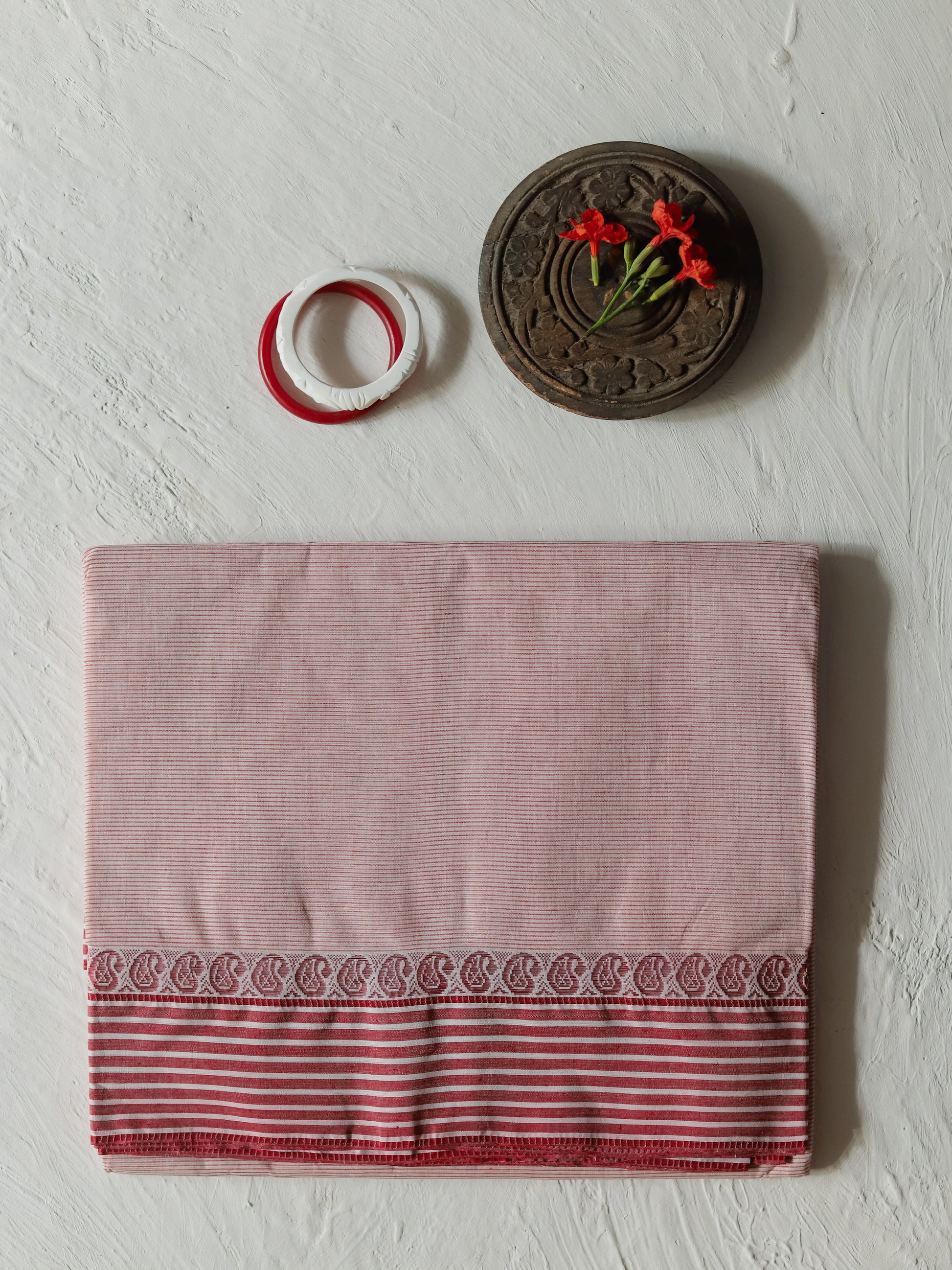 A folded Bengali white red cotton sari, featuring a red and white striped border with a paisley pattern, displayed on a textured off-white background with decorative props.