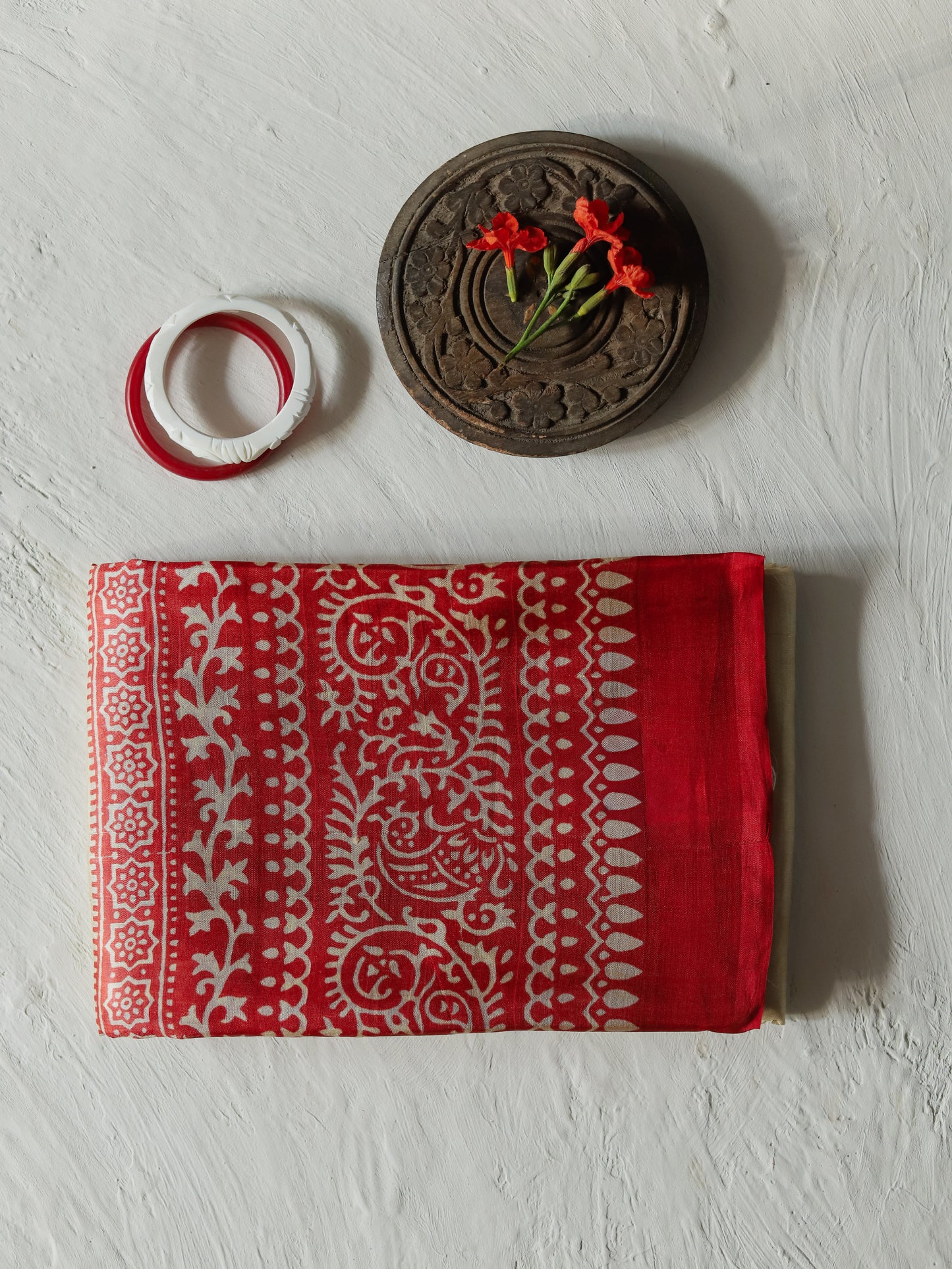 A folded handwoven white red mulberry silk saree  with a white traditional block-print pattern, displayed on a textured white background alongside a carved wooden decorative piece and red flower.
