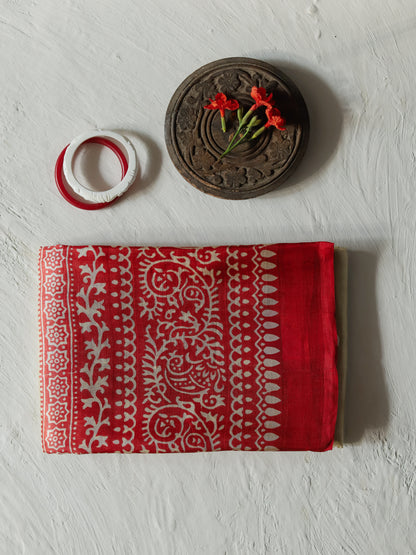 A folded handwoven white red mulberry silk saree  with a white traditional block-print pattern, displayed on a textured white background alongside a carved wooden decorative piece and red flower.