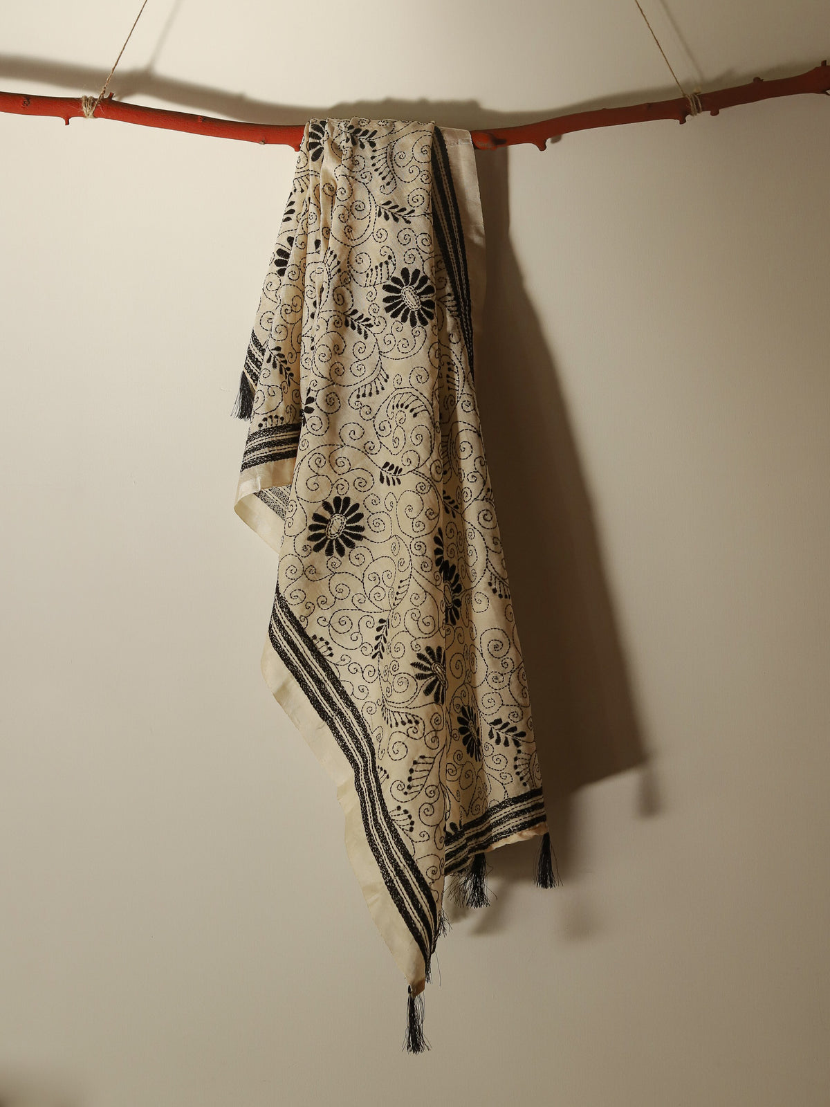A cream-colored Pobon Bengal Kantha Hand Embroidery Tussar Silk Stolewith black floral and scroll patterns, hanging from a rustic wooden branch against a plain beige wall.