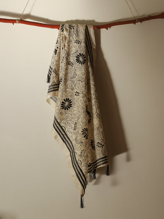 A cream-colored Pobon Bengal Kantha Hand Embroidery Tussar Silk Stolewith black floral and scroll patterns, hanging from a rustic wooden branch against a plain beige wall.