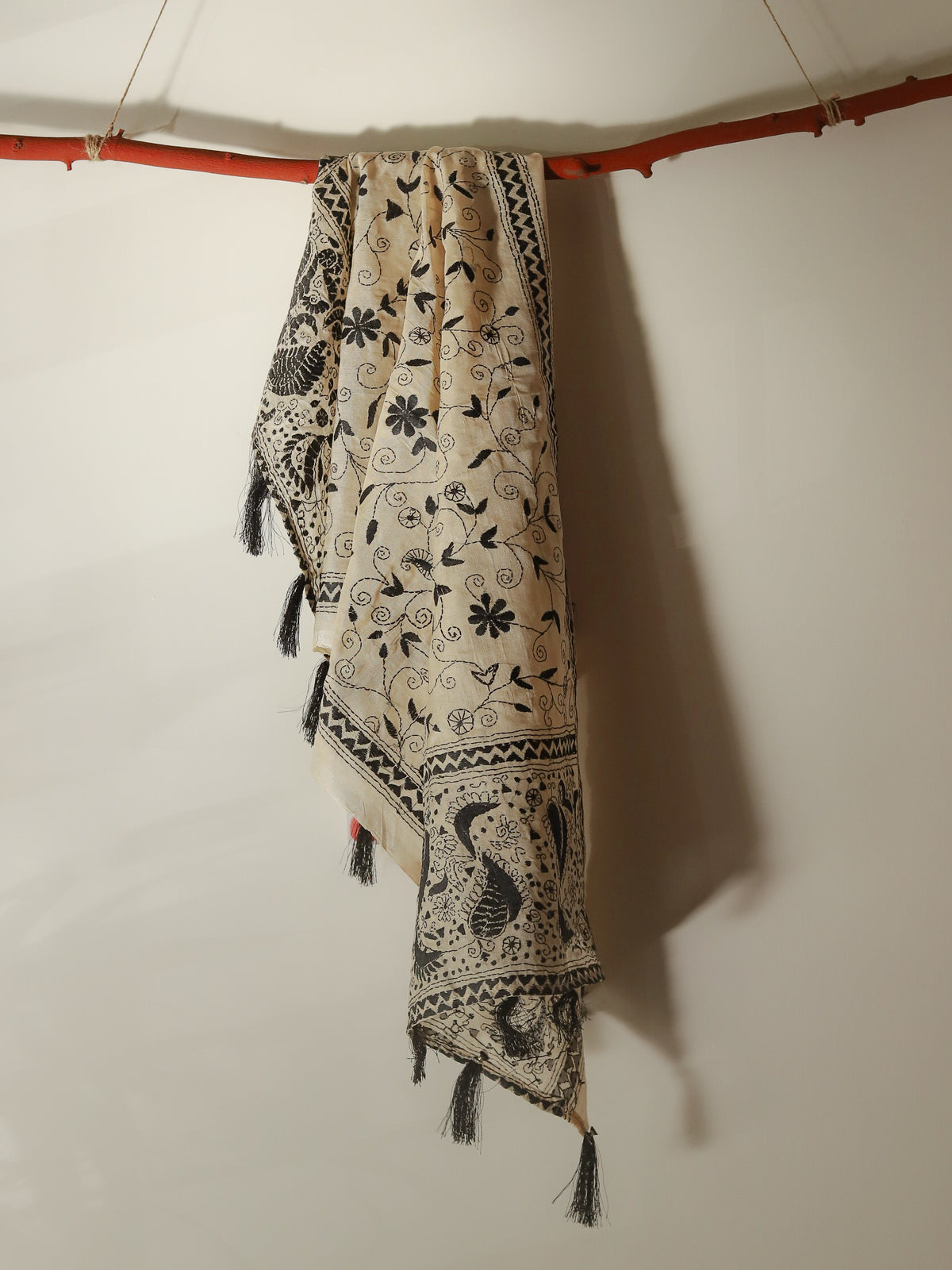 A cream-colored Pobon Bengal Kantha Hand Embroidery Tussar Silk Stole or scarf with black floral and geometric patterns, hanging from a rustic orange-brown wooden branch against a plain off-white wall.