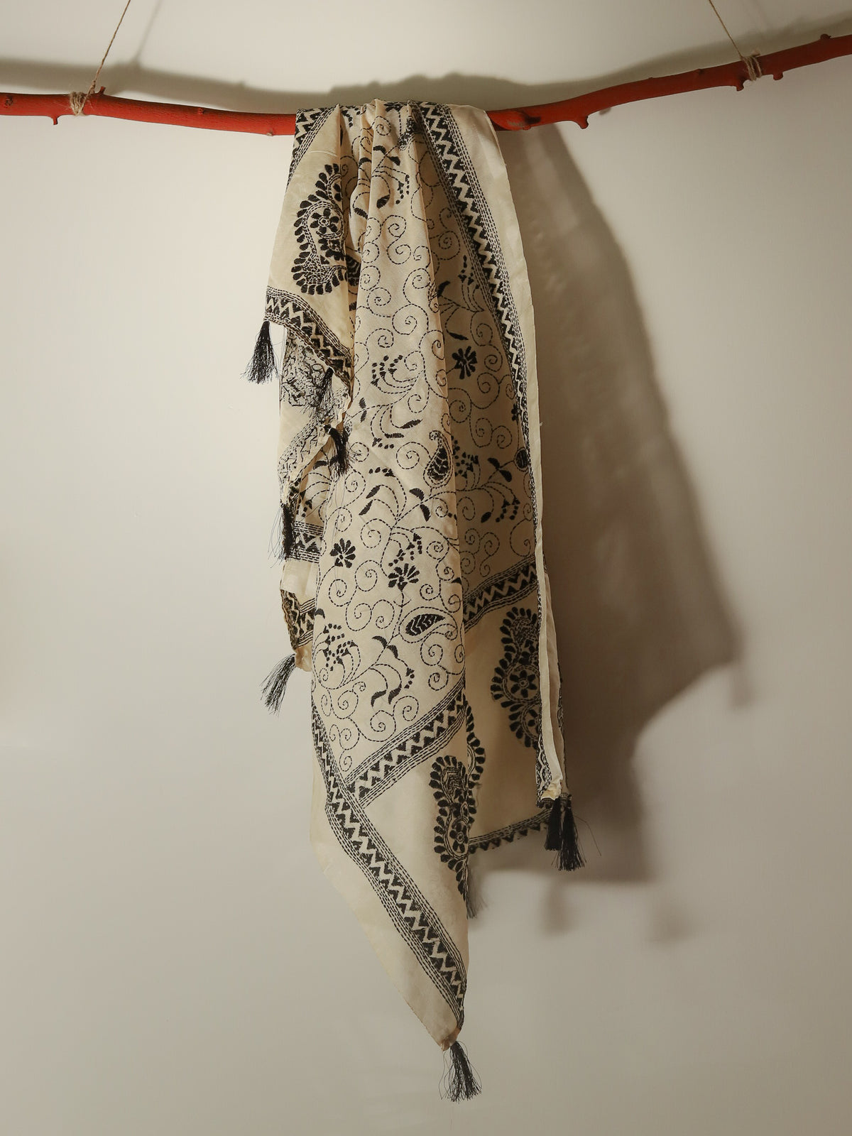 A cream-colored Pobon Bengal Kantha Hand Embroidery Tussar Silk Stole or scarf with black floral and scroll patterns, hanging from a rustic orange-brown wooden rod against a plain off-white wall.