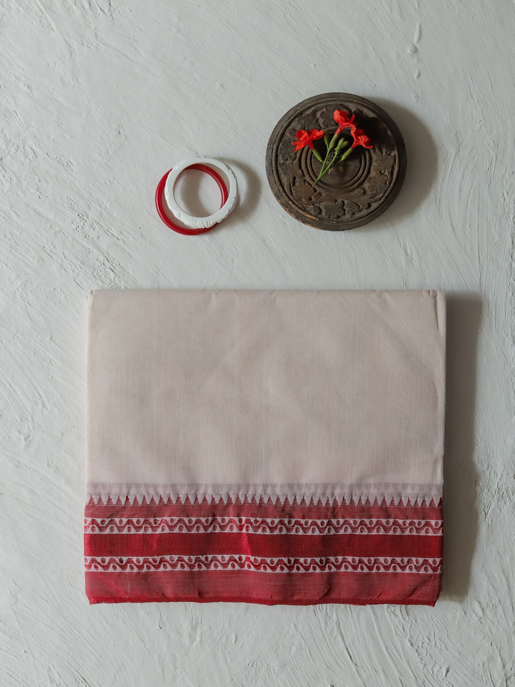 A folded Bengal handwoven red white cotton saree with a red and white patterned border at the bottom, displayed on a textured white wall alongside a small carved wooden ornament and two rings.