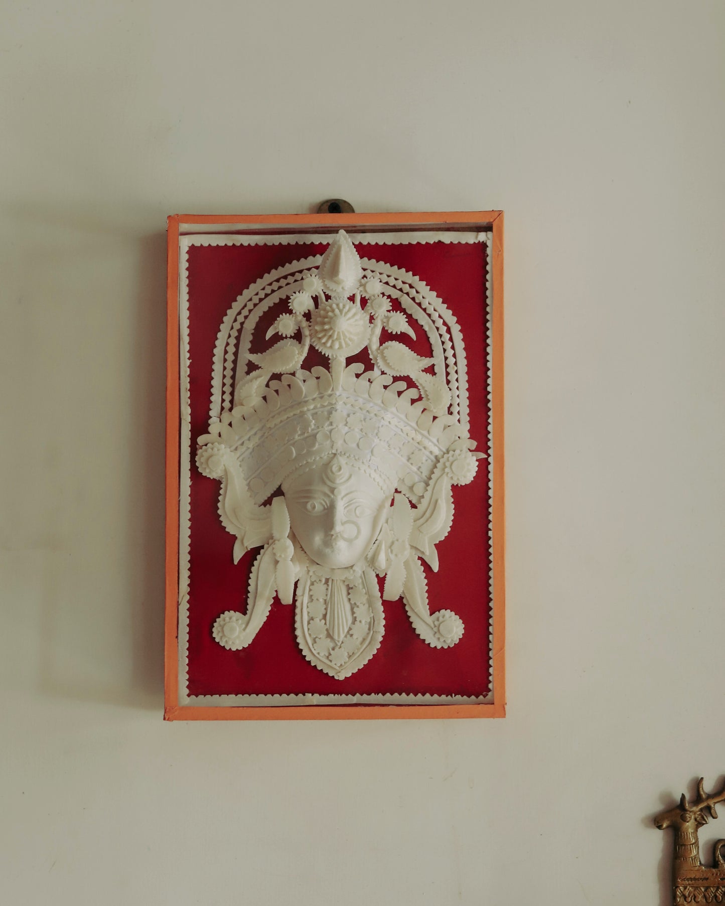 Decorative wall art with a Bengal handmade white sholapith face design on a red background, framed in glass.