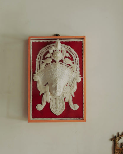 Decorative wall art with a Bengal handmade white sholapith face design on a red background, framed in glass.