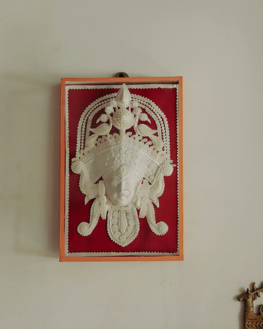 Decorative wall art with a Bengal handmade white sholapith face design on a red background, framed in glass.
