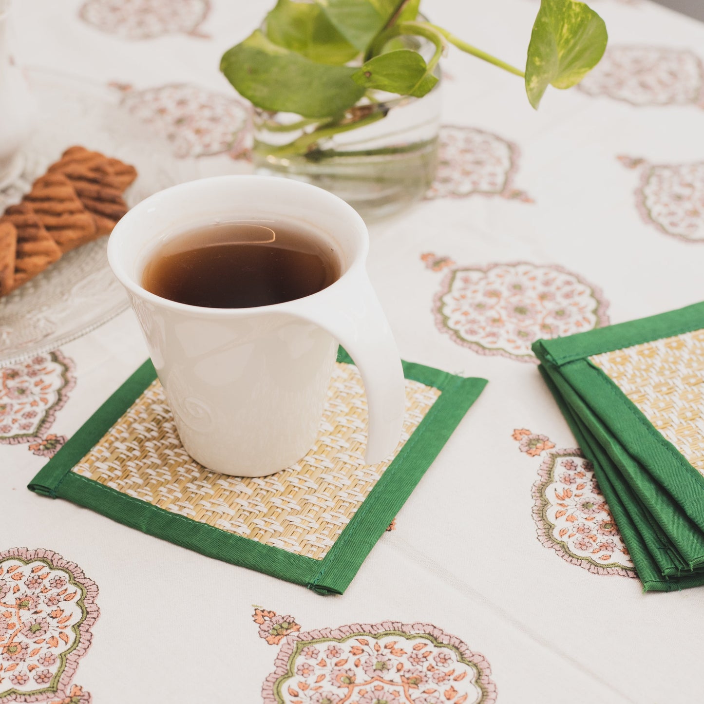 White cup of tea on a patterned tablecloth with green handwoven table coasters and cookies