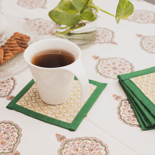 White cup of tea on a patterned tablecloth with green handwoven table coasters and cookies