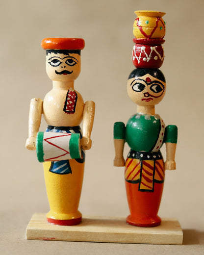 Two handcrafted wooden dolls, one representing a man and the other a woman, with colorful traditional attire. The man doll has a mustache and wears a yellow dhoti, while the woman doll wears a green and red saree with a multicolored pot on her head.