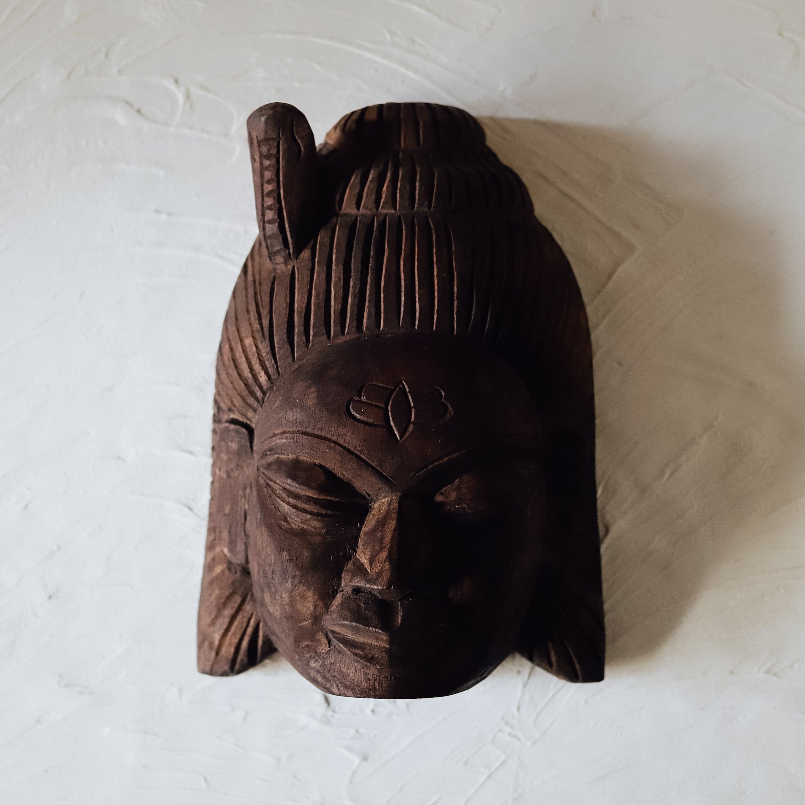 A hand-carved wooden mask with a traditional design, featuring intricate facial features, a forehead motif, and a hairstyle, displayed against a white background.