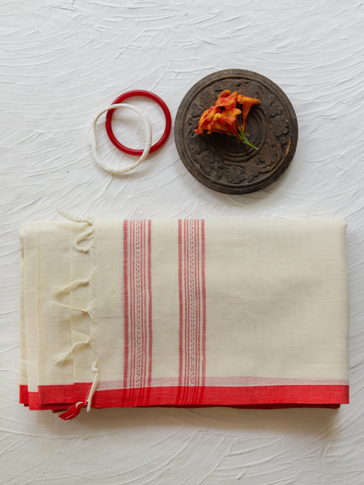 A handwoven Begumpuri cotton saree in red and white of West Bengal, displayed with a blouse piece, a red ring, and some dried flowers, on a white background.