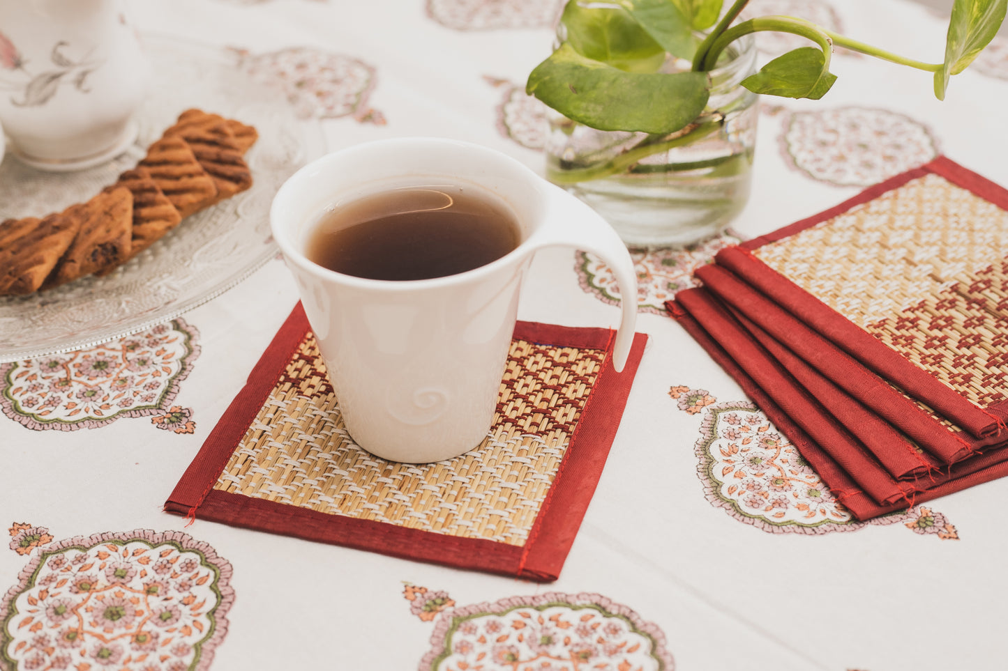 A set of six red and white patterned coasters displayed on a table with a cup of tea and a plate of cookies in the background.