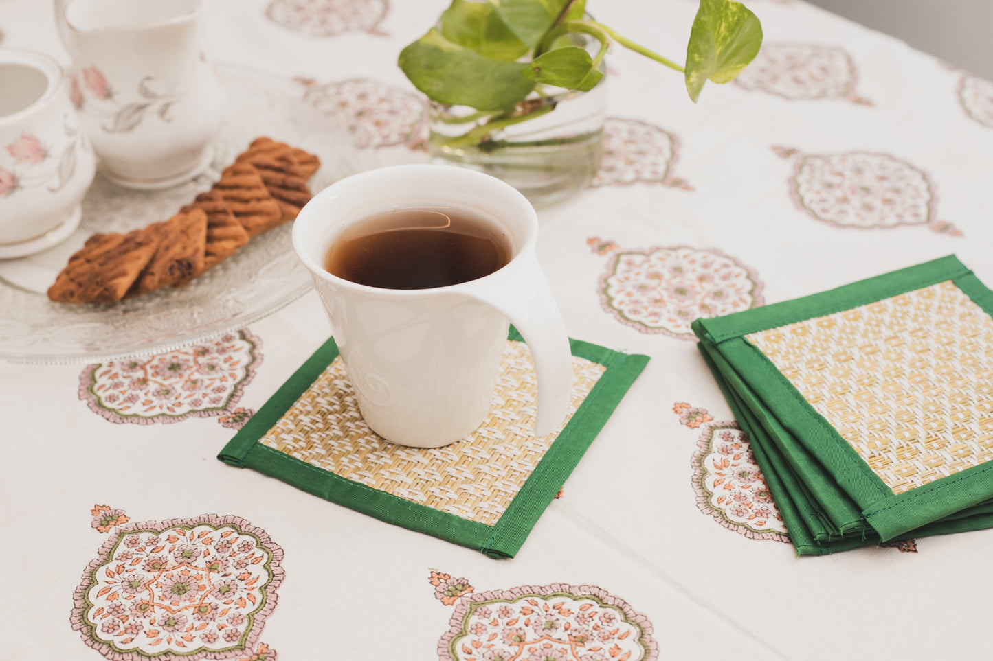 A set of handwoven green and white patterned coasters displayed on a table with a cup of tea and a cookie, surrounded by a floral-patterned tablecloth.