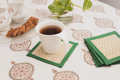 A set of handwoven green and white patterned coasters displayed on a table with a cup of tea and a cookie, surrounded by a floral-patterned tablecloth.